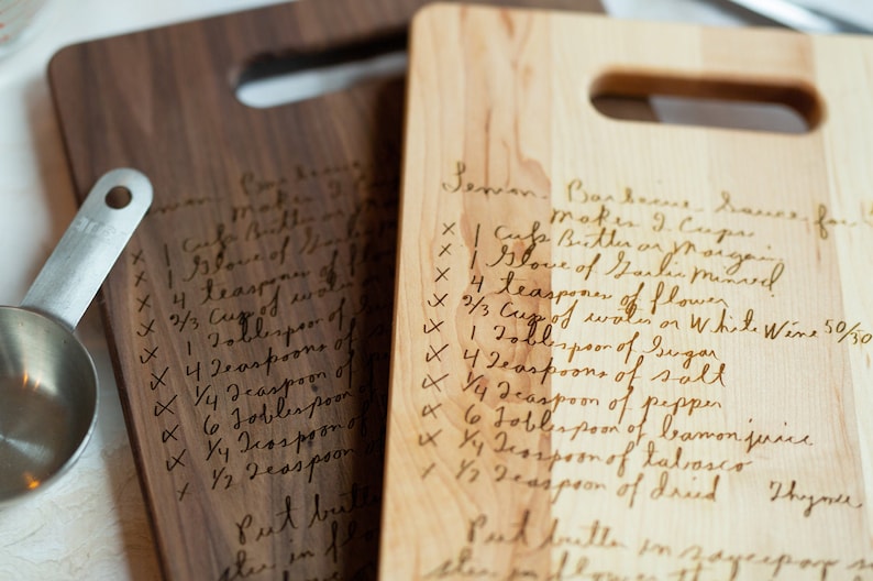 Pode incluir: Duas t&aacute;buas de corte de madeira com receitas manuscritas gravadas. Uma t&aacute;bua &eacute; marrom escuro, a outra marrom claro. Ambas t&ecirc;m uma al&ccedil;a. Um copo medidor &eacute; vis&iacute;vel &agrave; esquerda. As receitas est&atilde;o em it&aacute;lico.