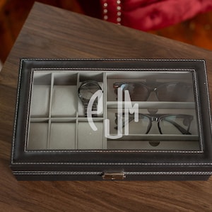 Puede incluir: Un organizador negro de polipiel para relojes y gafas con tapa de cristal transparente. El interior tiene compartimentos grises para relojes y gafas. Se ve un reloj negro y dos pares de gafas. Las iniciales "EJM" están grabadas en el cristal.