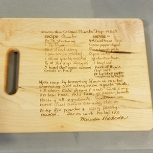 Pode incluir: Uma t&aacute;bua de corte de madeira clara com uma al&ccedil;a, gravada com uma receita de gumbo manuscrita. O texto inclui ingredientes e instru&ccedil;&otilde;es, com a assinatura do autor na parte inferior. Um item de cozinha pr&aacute;tico.