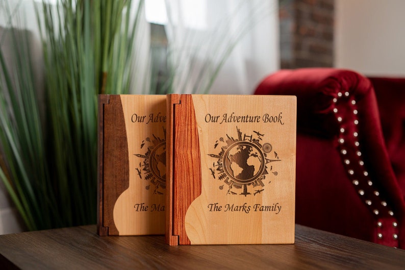 K&ouml;nnte beinhalten: Zwei h&ouml;lzerne Abenteuerb&uuml;cher mit eingravierten Weltkartenmotiven. Die B&uuml;cher tragen den Text "Our Adventure Book" und "The Marks Family" auf dem Einband. Die B&uuml;cher sind aus hellem und dunklem Holz gefertigt.