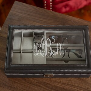 May include: A black leatherette glasses and watch organizer with a clear glass top. The interior is divided into compartments with gray lining. The organizer contains a watch, sunglasses, and eyeglasses. A monogram is etched on the glass.