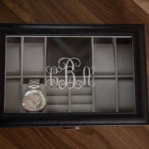 May include: Black leather watch box with a clear top, featuring a silver watch and a silver monogram. The interior is lined with gray fabric, providing storage for multiple watches and accessories. The box sits on a wooden surface.
