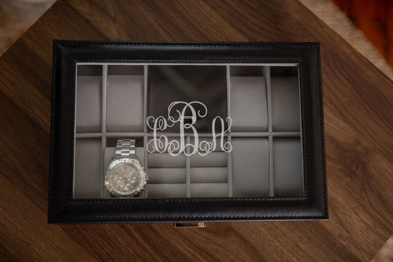 May include: Black leather watch box with a clear top, featuring a silver watch and a silver monogram. The interior is lined with gray fabric, providing storage for multiple watches and accessories. The box sits on a wooden surface.