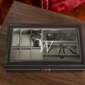 May include: Black leather watch and glasses organizer with a clear glass top. The organizer contains a black watch, sunglasses, and eyeglasses. A white letter "W" is visible on the glass. The organizer is on a wooden surface.