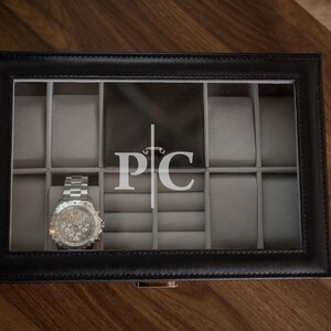Puede incluir: Caja de reloj de cuero negro con tapa transparente, que contiene un reloj de pulsera plateado. El interior es gris con compartimentos para relojes y anillos. Las iniciales "PC" se muestran en blanco en la parte posterior de la caja.