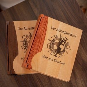 K&ouml;nnte beinhalten: Zwei h&ouml;lzerne Abenteuerb&uuml;cher mit hellen Holzdeckeln und dunklen Holzr&uuml;cken. Die Deckel sind mit einem Weltkarten-Design und dem Text "Our Adventure Book" und "Matt and Elizabeth" graviert. Die B&uuml;cher sind ge&ouml;ffnet.