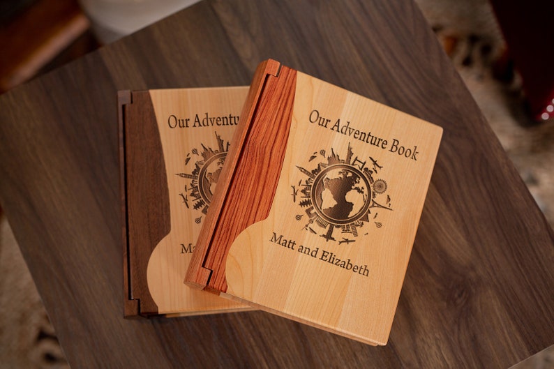 K&ouml;nnte beinhalten: Zwei h&ouml;lzerne Abenteuerb&uuml;cher mit hellen Holzdeckeln und dunklen Holzr&uuml;cken. Die Deckel sind mit einem Weltkarten-Design und dem Text "Our Adventure Book" und "Matt and Elizabeth" graviert. Die B&uuml;cher sind ge&ouml;ffnet.