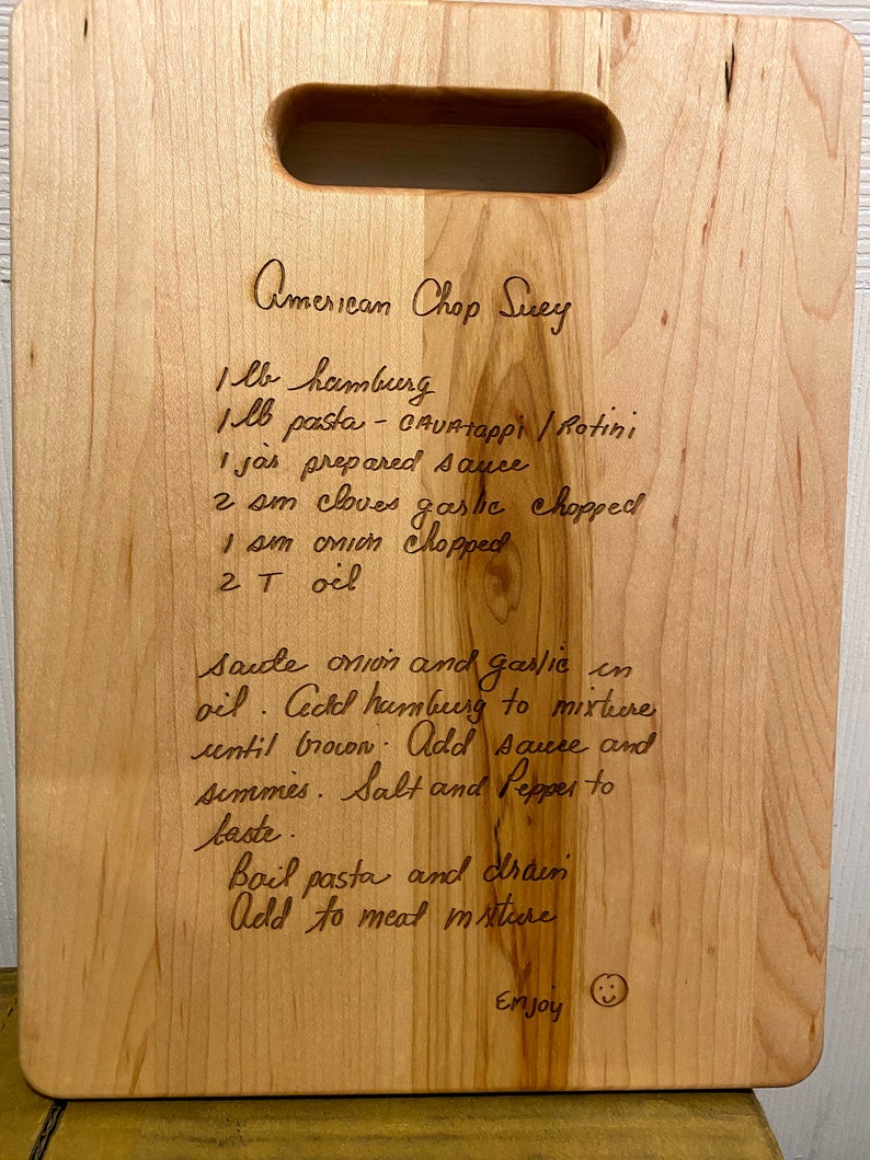 Pode incluir: T&aacute;bua de corte retangular de madeira clara com uma pega, com uma receita gravada de "American Chop Suey". A receita inclui ingredientes como carne picada e massa. A t&aacute;bua tem um gr&atilde;o de madeira claro e um pequeno smiley.