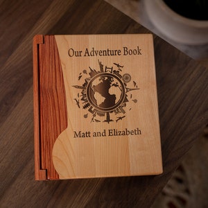 K&ouml;nnte beinhalten: Ein h&ouml;lzernes Abenteuerbuch mit hellem Holzdeckel und dunklerem Holzr&uuml;cken. Der Einband ist mit den Worten "Our Adventure Book", einem Globus und Reisesymbolen graviert. Die Namen "Matt and Elizabeth" sind ebenfalls eingraviert.