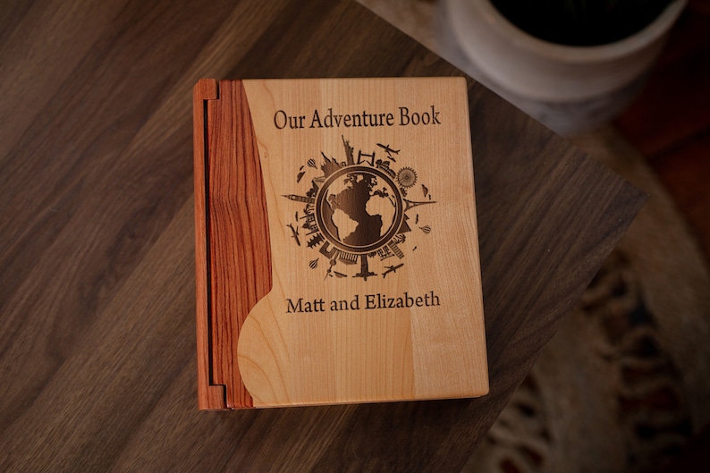 K&ouml;nnte beinhalten: Ein h&ouml;lzernes Abenteuerbuch mit hellem Holzdeckel und dunklerem Holzr&uuml;cken. Der Einband ist mit den Worten "Our Adventure Book", einem Globus und Reisesymbolen graviert. Die Namen "Matt and Elizabeth" sind ebenfalls eingraviert.