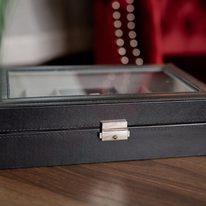 May include: A black watch box with a glass top and a silver clasp. The box is rectangular and appears to be made of leather or a leather-like material. The box is sitting on a wooden surface.