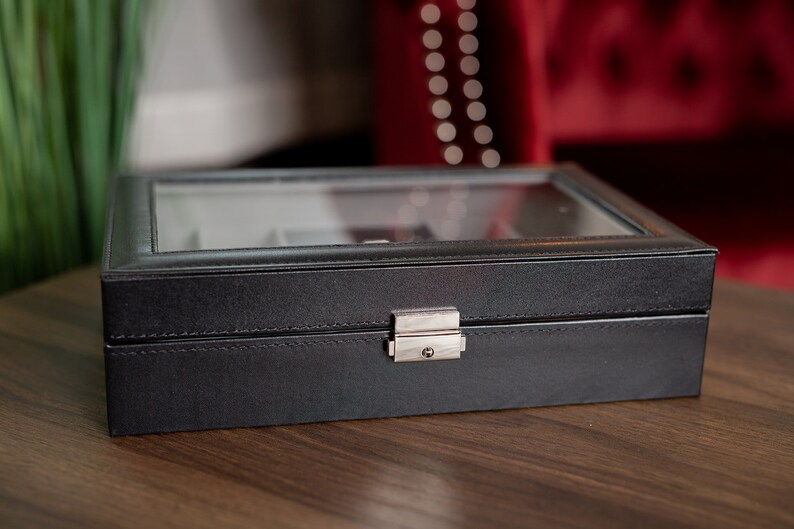 May include: A black watch box with a glass top and a silver clasp. The box is rectangular and appears to be made of leather or a leather-like material. The box is sitting on a wooden surface.
