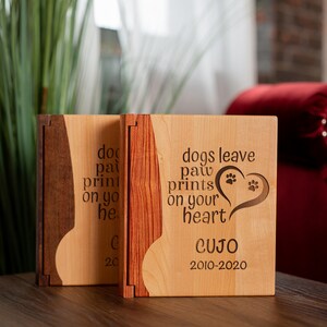May include: Two wooden memorial boxes with a light wood finish and dark wood accents. The boxes have the text "dogs leave paw prints on your heart" and the name "CUJO" with dates 2010-2020.