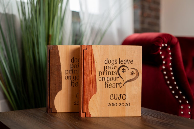 May include: Two wooden memorial boxes with a light wood finish and dark wood accents. The boxes have the text "dogs leave paw prints on your heart" and the name "CUJO" with dates 2010-2020.