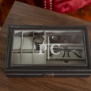 May include: Black leatherette watch and glasses organizer with a clear glass top. The organizer has compartments for a watch, sunglasses, and eyeglasses. The initials "PC" are displayed on the glass. The organizer is on a wooden surface.
