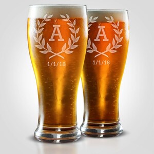 May include: Two tall beer glasses filled with golden beer and white foam. Each glass has a decorative design with the letter "A" inside a laurel wreath, and the date "1/1/18" below. The glasses are on a white background.