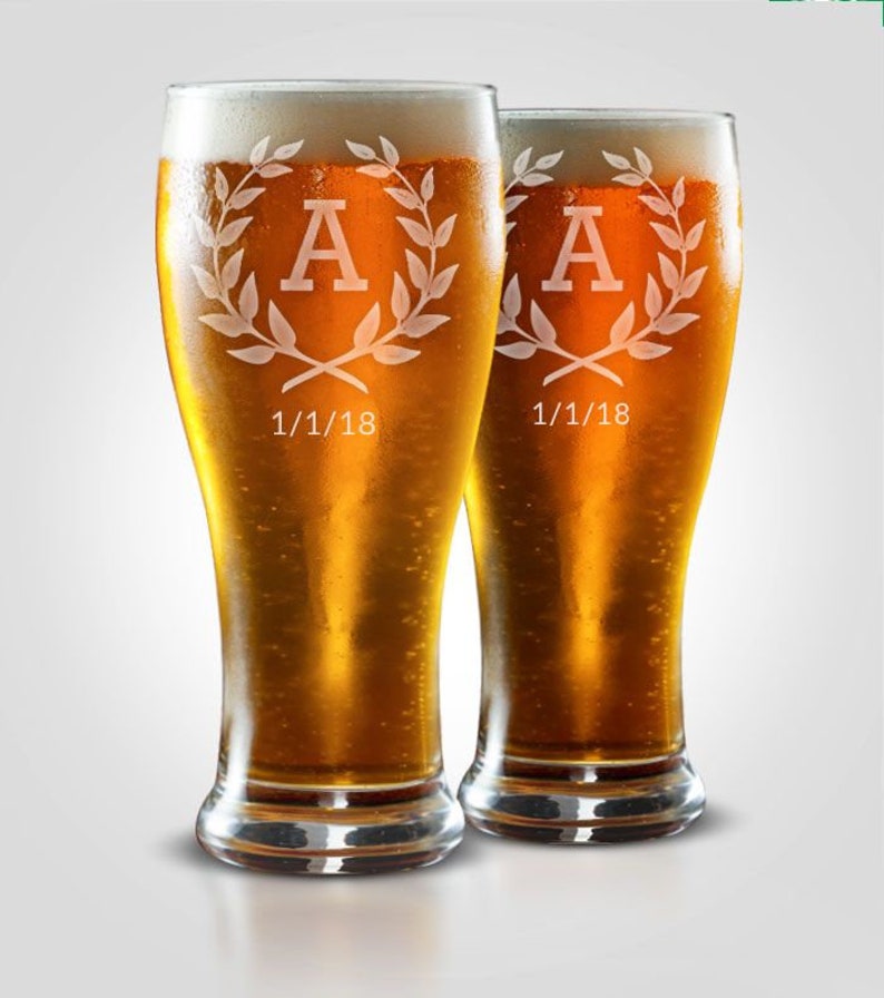 May include: Two tall beer glasses filled with golden beer and white foam. Each glass has a decorative design with the letter "A" inside a laurel wreath, and the date "1/1/18" below. The glasses are on a white background.