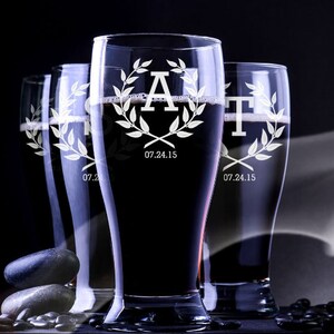 May include: Four tall, clear beer glasses filled with dark liquid. Each glass is engraved with a letter, a laurel wreath, and the date 07.24.15. The glasses are arranged in a row against a dark background, with a few dark stones in the foreground.
