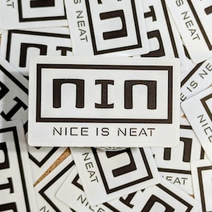 Puede incluir: Una tarjeta rectangular blanca con el texto "NIN" en negrita negra, con las palabras "NICE IS NEAT" debajo. La tarjeta está rodeada de otras tarjetas con la palabra "NEAT" y un cuadrado negro.