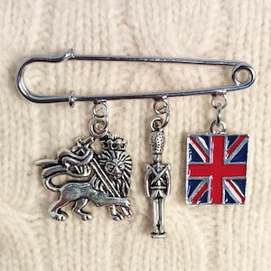 May include: A silver-toned safety pin brooch featuring three dangling charms: a lion holding a sword, a standing guard in a tall hat, and a rectangular charm displaying the red, white, and blue Union Jack flag design.