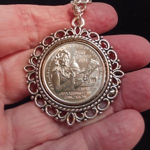 May include: A silver pendant necklace featuring a United States quarter coin with a portrait of Patsy Takemoto Mink, the first woman of color elected to the United States Congress. The coin reads "United States of America" and "25 Cents". The pendant is surrounded by a decorative silver frame.