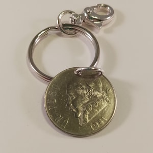 May include: A gold-colored coin keychain with a silver key ring and a silver clasp. The coin features a profile of a person with the text "UN PESO" and "1981" on it.