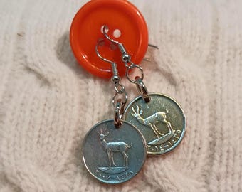 May include: Silver-toned dangle earrings featuring coin-shaped pendants with a deer design. The earrings are suspended from silver hooks and are attached to an orange button. The earrings are displayed on a textured, off-white surface.