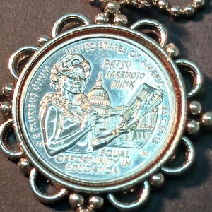 May include: A silver-colored pendant necklace featuring a U.S. quarter coin. The coin depicts Patsy Takemoto Mink, the U.S. Capitol building, and the text "Equal Opportunity in Education". The pendant has a decorative, openwork frame.