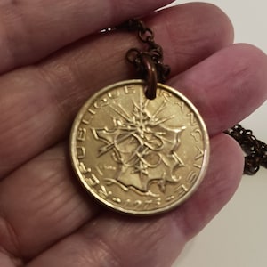 May include: A gold-colored coin pendant necklace with a French coin featuring the text "REPUBLIQUE FRANCAISE" and a stylized image of a tree. The coin is attached to a brown chain.