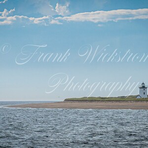 Pode incluir: Um farol branco com telhado preto fica em uma pequena ilha com grama verde e areia. O farol é cercado por água azul e um céu azul brilhante com nuvens brancas.