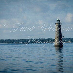 Op de afbeelding: Een stenen vuurtoren staat hoog in het midden van een kalm water. De lucht is lichtblauw met witte wolken. De vuurtoren is grijs met een groen dak.