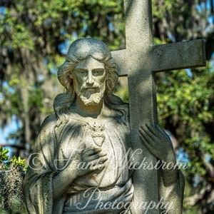 May include: A stone sculpture of Jesus Christ carrying a cross. He is wearing a robe and holding a heart in his left hand. The sculpture is in a cemetery setting with trees in the background.