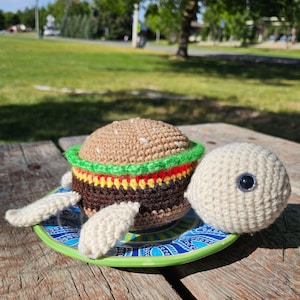 Könnte beinhalten: Eine gehäkelte Schildkröte hält einen gehäkelten Hamburger. Die Schildkröte ist weiß und der Hamburger ist braun mit grünem Salat, roter Tomate und gelbem Käse.