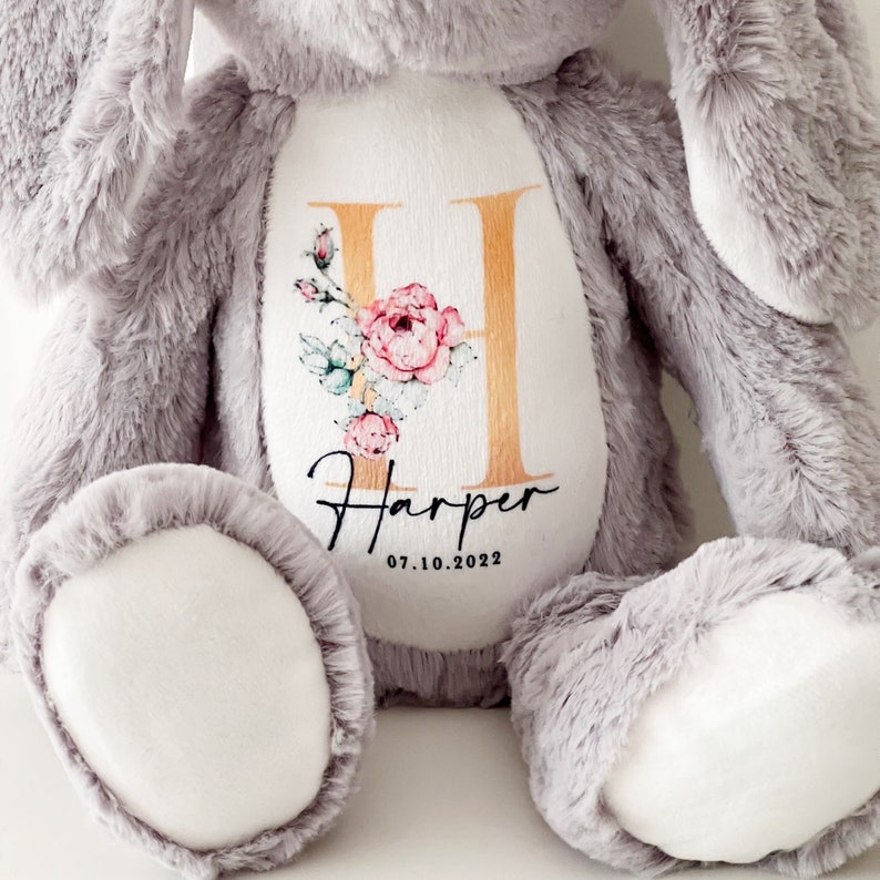 May include: A gray plush bunny with a white patch on its chest. The patch features a gold letter H with pink floral accents and the name "Harper" with the date "07.10.2022" underneath.
