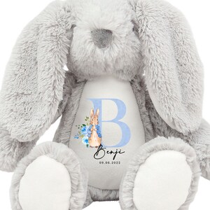 May include: A gray plush bunny with a white patch on its chest. The patch features a blue letter "B" with a watercolor illustration of Peter Rabbit and blue flowers. The name "Benji" and the date "09.06.2022" are written below the letter.