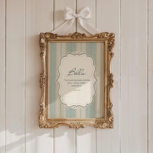 May include: A decorative framed print featuring the name "Bella". The gold-coloured ornate frame surrounds a print with a light blue and white striped pattern. The print includes the text "Tiny hands, big dreams, soft skies above. A heart so full, a world of love."