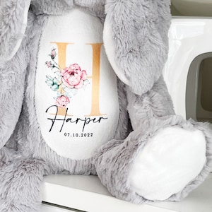 May include: A gray plush elephant with a white patch on its chest. The patch features a gold letter H with pink floral accents and the name "Harper" with the date "07.10.2022" underneath.