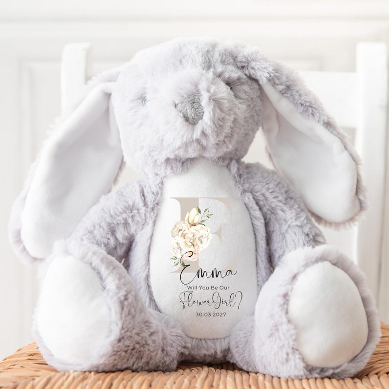 May include: A gray plush bunny with long ears and a white patch on its chest. The bunny has an embroidered letter "E" with floral accents and the text "Emma Will You Be Our Flower Girl? 30.03.2027".