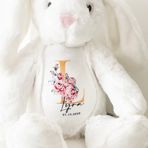 May include: White plush bunny with a gold letter L and pink floral design. The bunny has a personalized name "Lyra" and the date "07.10.2022" embroidered on the chest.