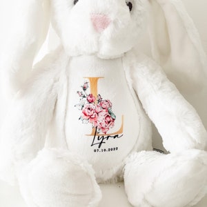May include: White plush bunny with a gold letter L and pink floral design. The bunny has a personalized name "Lyra" and the date "07.10.2022" embroidered on the chest.