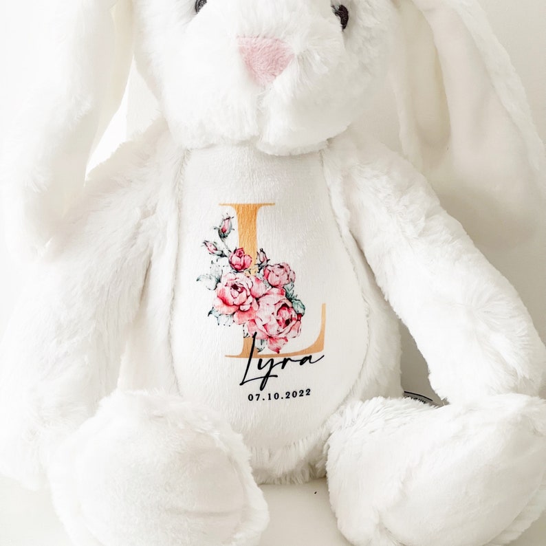 May include: White plush bunny with a gold letter L and pink floral design. The bunny has a personalized name "Lyra" and the date "07.10.2022" embroidered on the chest.