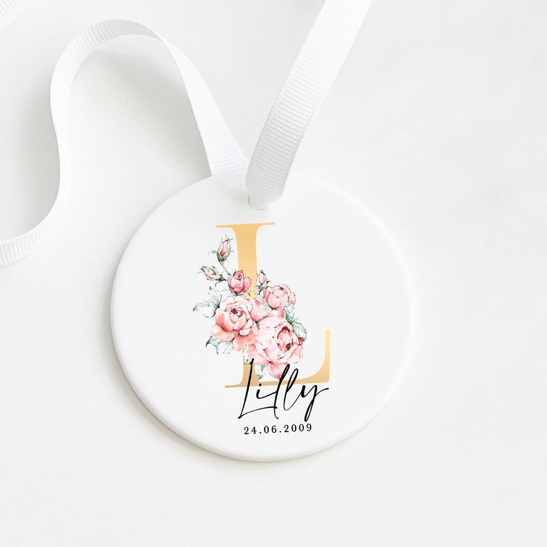 May include: A white ceramic ornament with a gold letter L and pink floral design. The ornament is decorated with the name "Lilly" and the date "24.06.2009".