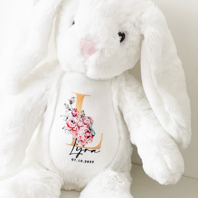 May include: White plush bunny with a gold letter L and pink floral design. The bunny has a personalized name "Lyra" and the date "07.10.2022" embroidered on the chest.