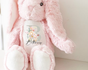 May include: Pink plush bunny with a white patch on its chest. The patch features a floral design with the letter "B" and the name "Bethany" written in cursive.
