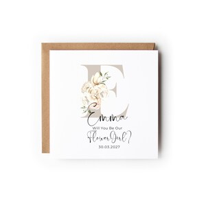 May include: A white card with a floral design and the letter E. The text on the card reads "Emma Will You Be Our Flower Girl? 30.03.2027"