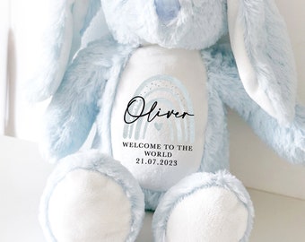 May include: A light blue plush bunny with long ears. The bunny has a white patch on its chest with a rainbow design and the text "Oliver, Welcome to the World, 21.07.2023".
