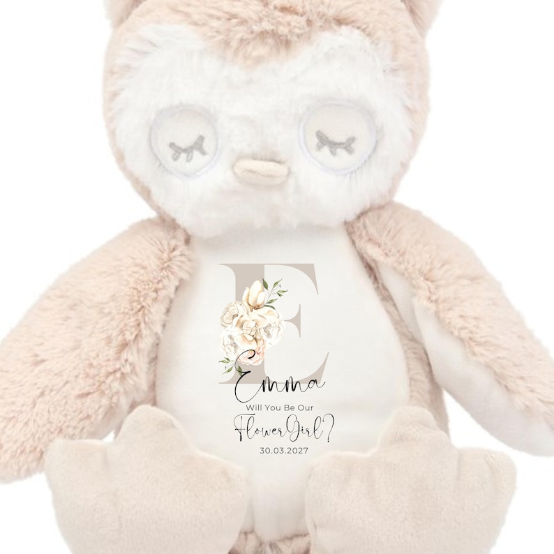 May include: A plush owl toy with a white and beige color scheme. The owl has a white face with closed eyes and a beige body. The owl is holding a white sign with the text "Emma Will You Be Our Flower Girl? 30.03.2022" and a floral letter E.