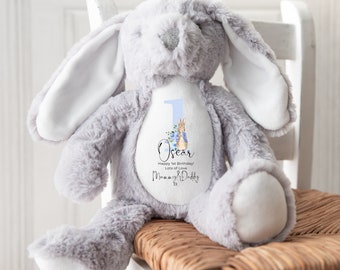 Op de afbeelding: Een zachtgrijs konijn met lange oren, zittend op een rieten stoel. Het konijn heeft een witte vlek op de borst met het nummer 1, een Peter Rabbit-illustratie en de naam "Oscar" met een verjaardagsbericht.