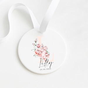 May include: A white ceramic ornament with a pink floral design and the letter L. The ornament also has the name "Lilly" and the date "24.06.2009" written in black.