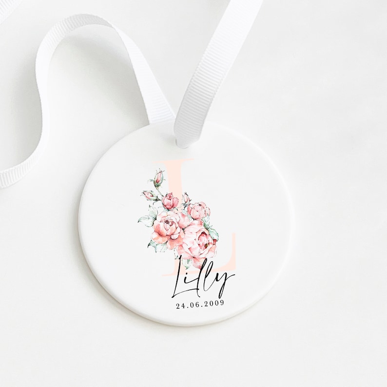 May include: A white ceramic ornament with a pink floral design and the letter L. The ornament also has the name "Lilly" and the date "24.06.2009" written in black.